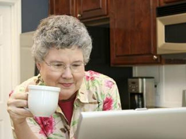 A senior woman sits in her kitchen holding a coffee cup in her right hand as she reads on her laptop computer.